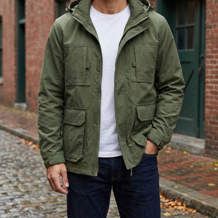 Man wearing the Pavia Hooded Cargo Jacket in Army Green open over a white tee on a brick-lined street