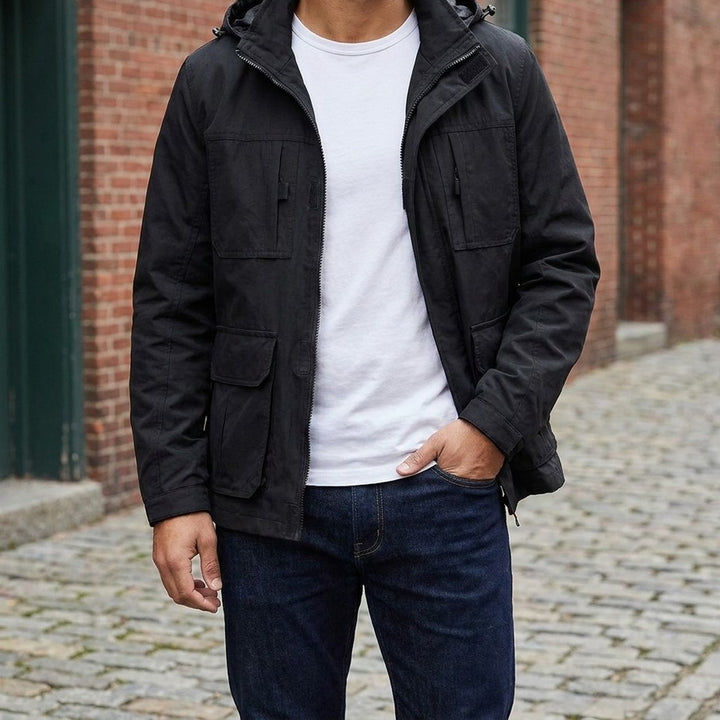 Man wearing the Pavia Hooded Cargo Jacket in Black open over a white tee on a brick-lined street
