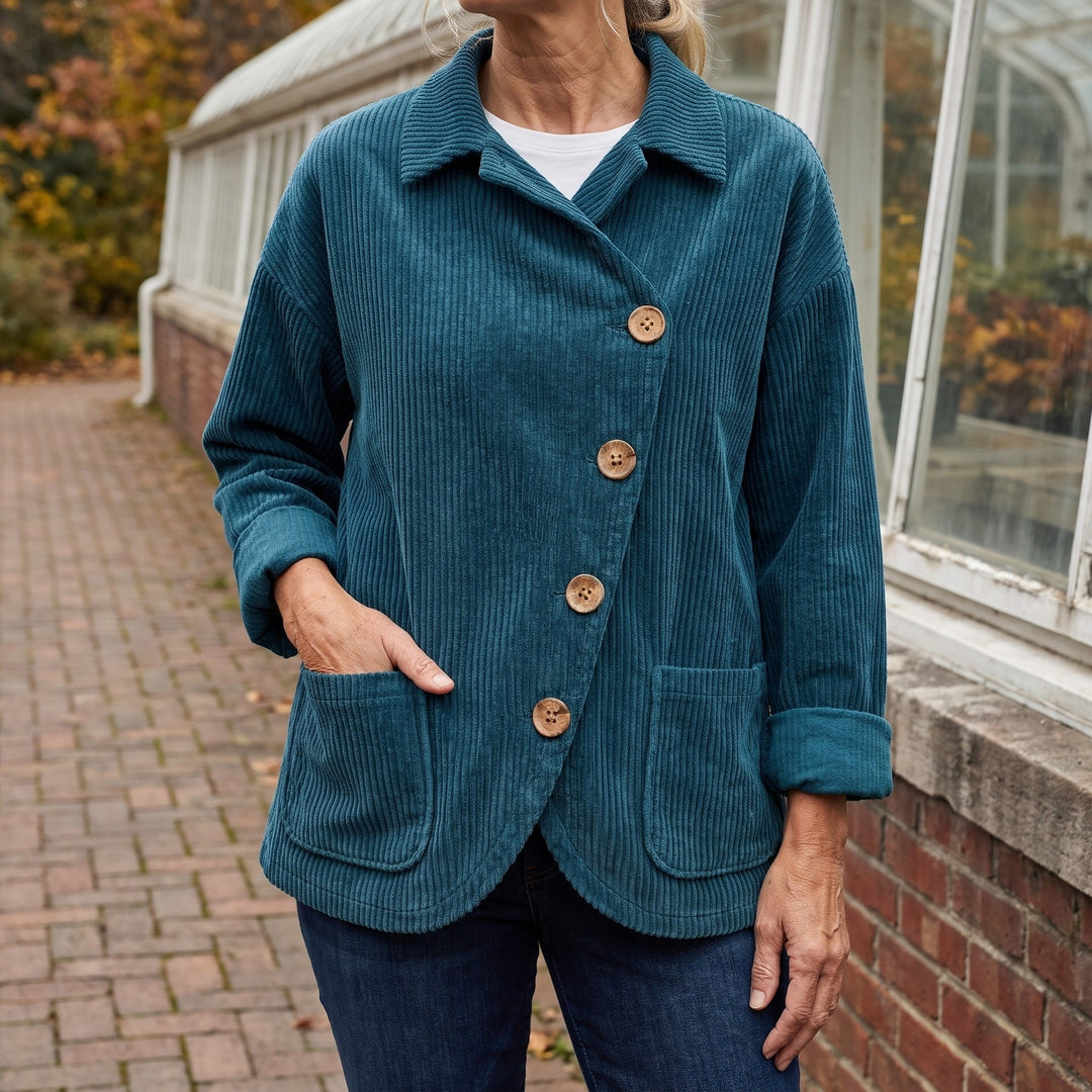 Asiago Corduroy Jacket — Blue, worn by a woman with silver-blonde hair over a white tee and dark jeans, one hand in the pocket, cropped at the neck, outside a glasshouse on a brick path