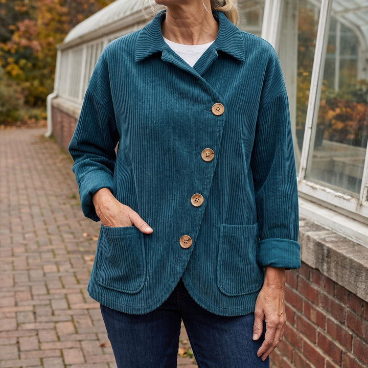 Asiago Corduroy Jacket — Blue, worn by a woman with silver-blonde hair over a white tee and dark jeans, one hand in the pocket, cropped at the neck, outside a glasshouse on a brick path