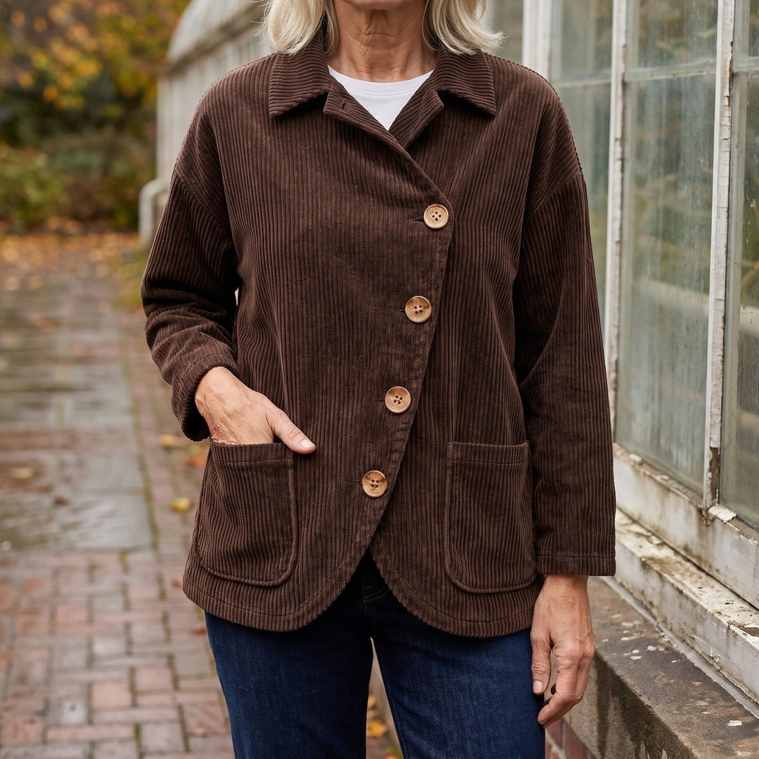 Asiago Corduroy Jacket — Brown, worn by a woman with silver-blonde hair over a white tee and dark jeans, one hand in the pocket, cropped at the neck, outside a glasshouse on a brick path