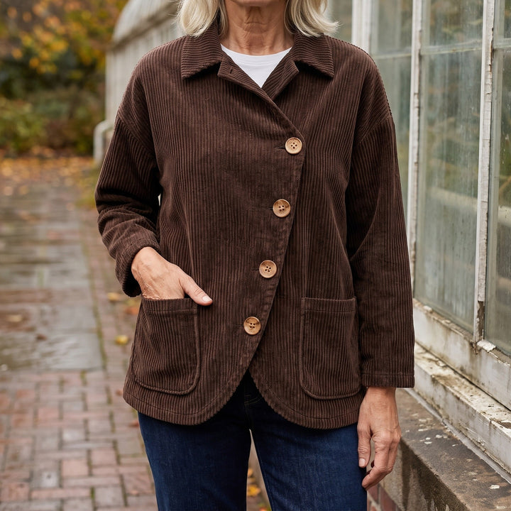 Asiago Corduroy Jacket — Brown, worn by a woman with silver-blonde hair over a white tee and dark jeans, one hand in the pocket, cropped at the neck, outside a glasshouse on a brick path