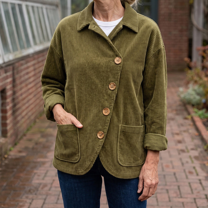 Asiago Corduroy Jacket — Olive, worn by a woman with silver-blonde hair over a white tee and dark jeans, one hand in the pocket, cropped at the neck, outside a glasshouse on a brick path