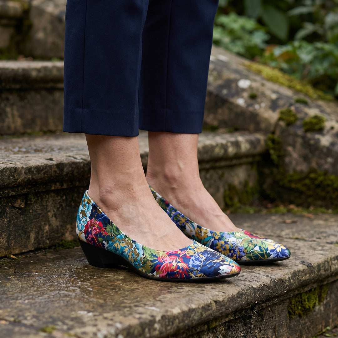 Camogli Floral Pumps — Teal Floral, worn by a woman in navy cropped trousers, knee down, on stone garden steps