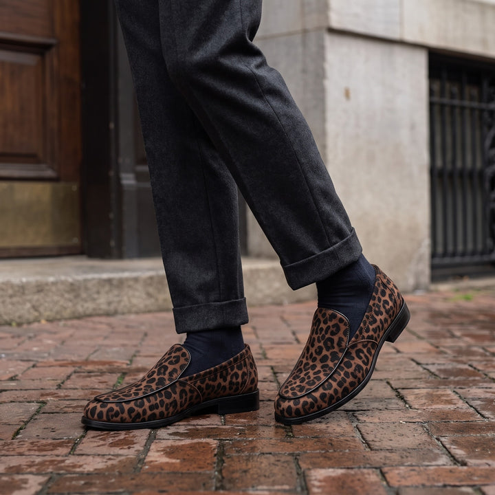 Polignano Leopard Loafers — Brown Leopard, worn by a man in charcoal trousers, knee down, on a brick sidewalk outside a historic hotel entrance